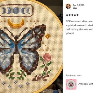 K&ouml;nnte beinhalten: Fertige Kreuzstichstickerei in einem Holzrahmen. Das Design zeigt einen blauen Schmetterling, einen Mond und Mondphasen. Rosa Blumen und Sterne umgeben den Schmetterling. Der Text "Antisocial Butterfly Cross Stitch Pattern" ist sichtbar.