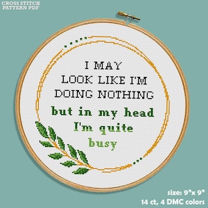 Op de afbeelding: Een kruissteekpatroon voor een hoepel met een groene bladertak en een gouden cirkel met de tekst "I MAY LOOK LIKE I'M DOING NOTHING but in my head I'm quite busy". De hoepel is 9 inch bij 9 inch en gebruikt 14 count stof met 4 DMC-kleuren.