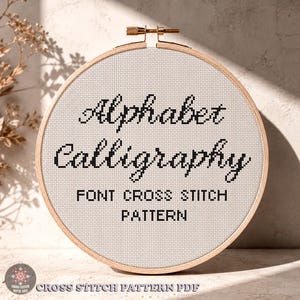 May include: A circular wooden embroidery hoop containing white Aida fabric with black cross-stitched text that reads Alphabet Calligraphy FONT CROSS STITCH PATTERN. The design features a cursive script for the first two words and block letters below.