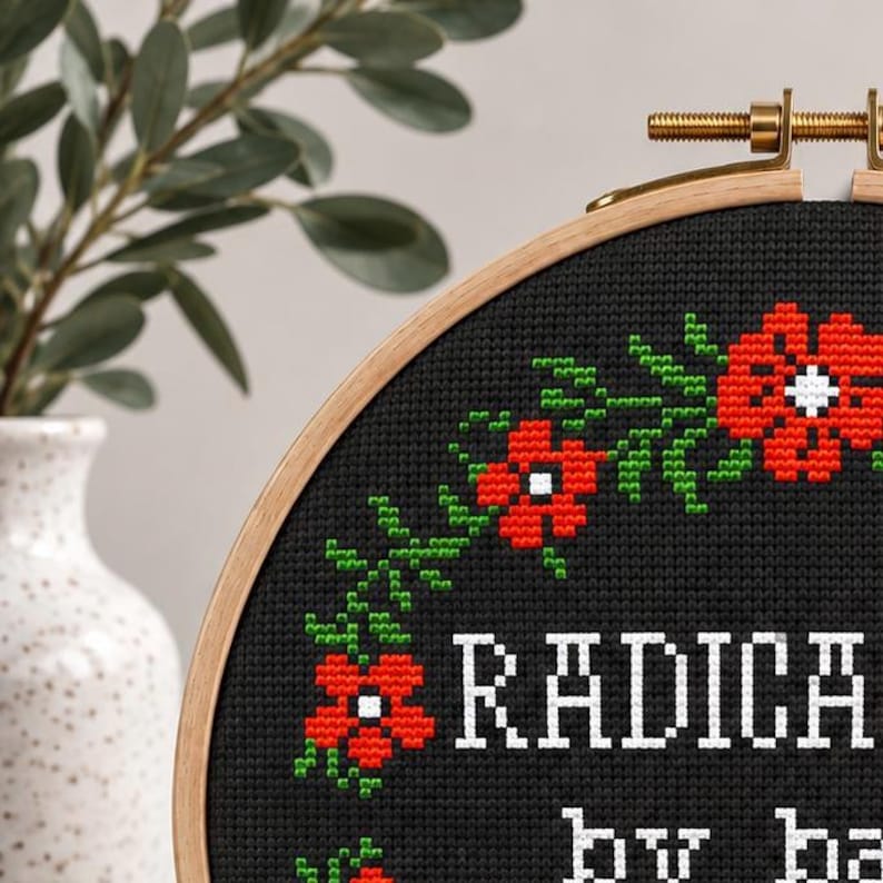 A cross-stitch pattern PDF with the words "RADICALIZED by basic decency" in white. The design is surrounded by a wreath of red flowers and green leaves. The hoop is set against a wooden background with floral accents.