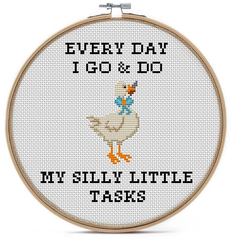 May include: A circular wooden embroidery hoop containing white fabric with a cross-stitch design of a white goose with an orange beak and feet, wearing a blue bow tie. The black text reads: EVERY DAY I GO & DO MY SILLY LITTLE TASKS.