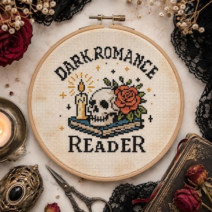 Könnte beinhalten: Ein runder Stickrahmen aus Holz mit einem Kreuzstichmotiv auf cremefarbenem Stoff. Das Design zeigt einen Totenkopf, eine rote Rose und eine brennende Kerze auf einem Buch, mit dem schwarzen Text "DARK ROMANCE READER" darüber und darunter.