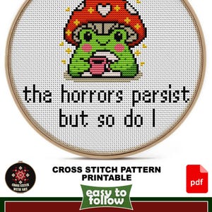 May include: Cross-stitch pattern in a wooden hoop featuring a green frog wearing a mushroom hat, holding a pink cup. The text reads "the horrors persist but so do I." Design size is 6" x 5".
