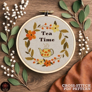 以下が含まれることがあります： 「Tea Time」の文字とティーポットのデザインが施されたクロスステッチ刺繍枠。オレンジと黄色の花と緑の葉が特徴です。背景は薄い灰色の生地で、枠は木製です。