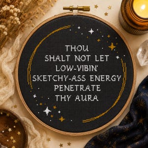 May include: A circular wooden embroidery hoop containing black fabric with white cross-stitched text that reads: THOU SHALT NOT LET LOW-VIBIN' SKETCHY-ASS ENERGY PENETRATE THY AURA. The design includes a gold stitched crescent moon and small white and gold stars.
