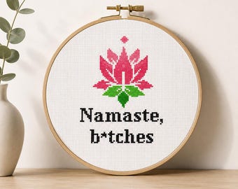 Puede incluir: Un bastidor de bordado circular de madera que contiene tela blanca con un diseño de punto de cruz. El diseño presenta una flor de loto rosa y verde sobre el texto negro que dice Namaste, b*tches, centrado en la tela dentro del marco.