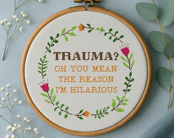 May include: A cross-stitch embroidery hoop with the text "TRAUMA? OH YOU MEAN THE REASON I'M HILARIOUS" in brown thread. The design is surrounded by a green and pink floral wreath. The wooden hoop is displayed on a light blue surface.