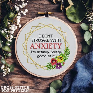 Könnte beinhalten: Ein Kreuzstich-Stickrahmen mit dem Text "I DON'T STRUGGLE WITH ANXIETY, I'm actually pretty good at it." Das Design umfasst einen goldenen geometrischen Rahmen und florale Akzente in Rot, Gelb und Lila. Die Worte "CROSS-STITCH PDF PATTERN" stehen unten.