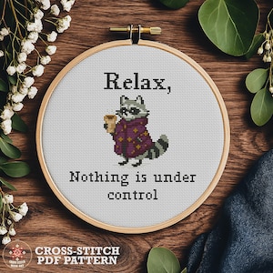 May include: Cross-stitch PDF pattern in a wooden hoop. The design features a raccoon holding a coffee cup, wearing a purple robe, and the text "Relax, Nothing is under control."