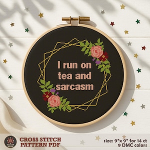 May include: A black cross-stitch embroidery hoop with the phrase "I run on tea and sarcasm" in pink. The design includes a gold geometric frame and floral accents in pink, red, and green. The hoop is 9 inches in diameter.