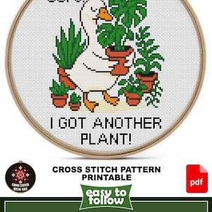 May include: Cross-stitch pattern featuring a white goose with orange beak and feet, holding potted plants. Text reads "OOPS! I GOT ANOTHER PLANT!" in black. The design is in a wooden embroidery hoop.