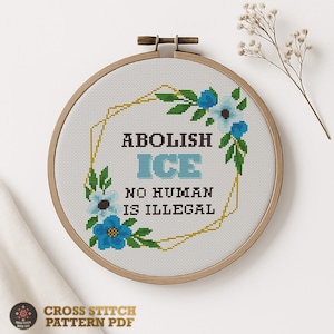 Op de afbeelding: Een kruissteekborduurwerk in een houten hoepel toont de tekst "ABOLISH ICE, NO HUMAN IS ILLEGAL". Het ontwerp bevat blauwe bloemen, groene bladeren en een gouden geometrische rand. De woorden "CROSS STITCH PATTERN PDF" staan onderaan.