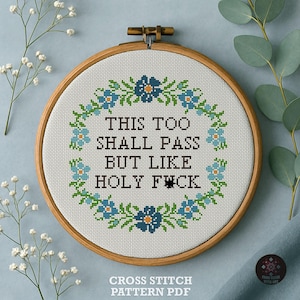 Puede incluir: Un bordado de punto de cruz en un aro de madera presenta el texto "THIS TOO SHALL PASS BUT LIKE HOLY F*CK" rodeado por una corona floral. El diseño incluye elementos florales azules y verdes sobre un fondo blanco.