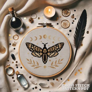 May include: Cross stitch pattern PDF featuring a black and white moth with gold accents, surrounded by a crescent moon design. The pattern is displayed in a wooden embroidery hoop on a white fabric background.