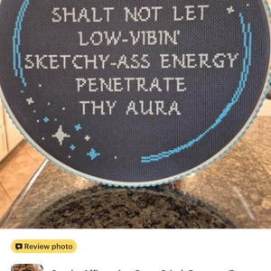 May include: A circular cross-stitch embroidery piece featuring a dark gray fabric background with white text that reads: THOU SHALT NOT LET LOW-VIBIN' SKETCHY-ASS ENERGY PENETRATE THY AURA. The design includes light blue stitched stars and a curved border.