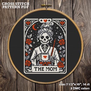 Cross-stitch pattern PDF featuring a skeletal figure holding a mug with a heart, surrounded by roses and stars. The design is framed in a wooden hoop, with the text "THE MOM" at the bottom. The pattern measures 7.5" x 10".
