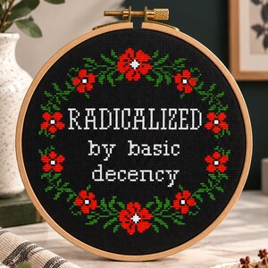A cross-stitch pattern PDF with the words "RADICALIZED by basic decency" in white. The design is surrounded by a wreath of red flowers and green leaves. The hoop is set against a wooden background with floral accents.