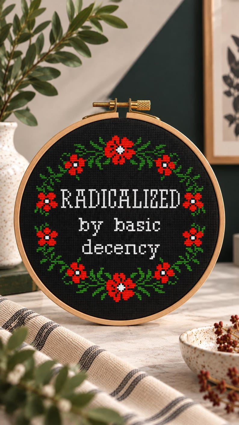 A cross-stitch pattern PDF with the words "RADICALIZED by basic decency" in white. The design is surrounded by a wreath of red flowers and green leaves. The hoop is set against a wooden background with floral accents.