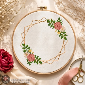 Può includere: Uno schema a punto croce con una cornice geometrica color oro decorata con rose rosa, piccoli fiori bianchi e foglie verdi su tessuto bianco, fissato in un telaio da ricamo circolare in legno con una vite di serraggio in metallo in alto.