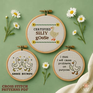 May include: Three cross-stitch patterns in wooden hoops featuring goose designs and funny phrases. The phrases include "Certified Silly Goose," "Goose Bumps," and "I think I will cause problems on purpose." Daisies are arranged around the hoops.