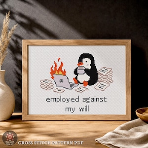 Op de afbeelding: Een ingelijst kruissteekpatroon met een zwart-witte pinguïn die een mok vasthoudt naast een brandende laptop en stapels papier. De tekst eronder luidt employed against my will. Het ontwerp gebruikt zwarte, witte, oranje en grijze garenkleuren.