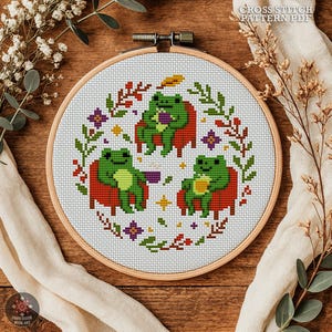 Puede incluir: Un patrón de punto de cruz en un aro de madera presenta tres ranas verdes sentadas en sillas rojas. El diseño está rodeado de motivos florales y de hojas en varios colores. El texto "CROSS STITCH PATTERN PDF" está en la parte superior.