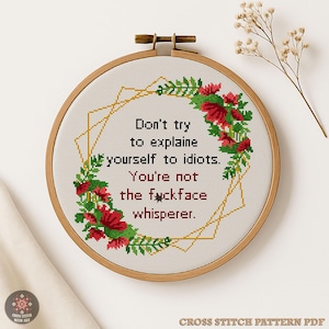Peut inclure: Une broderie au point de croix dans un cercle en bois affiche le texte "Don't try to explain yourself to idiots. You're not the fuckface whisperer." Le motif comprend une bordure florale avec des fleurs rouges et des feuilles vertes, et une forme géométrique dorée.