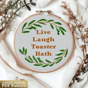 May include: A circular wooden embroidery hoop containing white Aida fabric with a cross-stitch design. The design features a green leaf wreath surrounding the brown text "Live Laugh Toaster Bath" centred in the middle of the fabric.