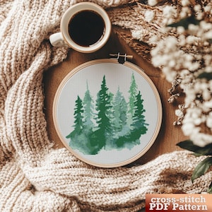 Peut inclure: Un motif de point de croix représentant une forêt avec des arbres verts, dans un style aquarelle. Le motif est présenté dans un tambour à broder en bois sur une couverture en tricot beige.