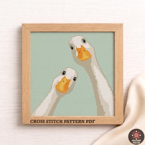 May include: A framed cross-stitch pattern PDF featuring two white geese with orange beaks and black eyes. The geese are set against a light green background. The text "CROSS STITCH PATTERN PDF" is at the bottom of the frame.