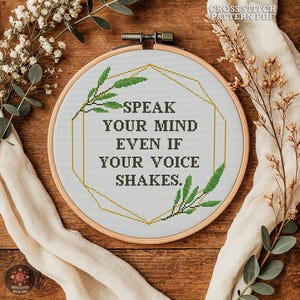 Op de afbeelding: Een kruissteek borduurring met de tekst "SPEAK YOUR MIND EVEN IF YOUR VOICE SHAKES." Het ontwerp heeft een geometrische gouden omtrek en groene bladaccenten. De woorden zijn donkergroen. De achtergrond is wit.