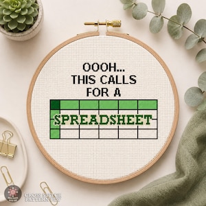 May include: A circular wooden embroidery hoop containing white Aida fabric with black cross-stitched text that reads OOOH... THIS CALLS FOR A SPREADSHEET, featuring a green and white grid pattern below the text representing a spreadsheet.