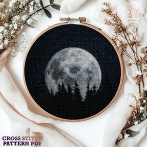 May include: A cross-stitch pattern in a wooden hoop depicts a full moon over a forest silhouette against a starry night sky. The text "CROSS STITCH PATTERN PDF" is at the bottom. Decorative flowers and fabric surround the hoop.