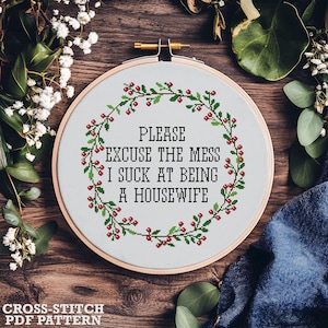 May include: A cross-stitch PDF pattern displayed in a wooden hoop. The design includes the text "PLEASE EXCUSE THE MESS I SUCK AT BEING A HOUSEWIFE" within a green and red wreath. The words "CROSS-STITCH PDF PATTERN" are at the bottom.