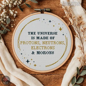 May include: A cross-stitch embroidery hoop with the text "THE UNIVERSE IS MADE OF PROTONS, NEUTRONS, ELECTRONS & MORONS". The design features gold and navy blue lettering and star accents. The hoop is set against a wooden background.