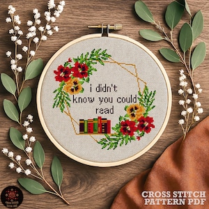 May include: A cross-stitch embroidery hoop with the text "I didn't know you could read." The design features a floral wreath with red and yellow flowers, green leaves, and a geometric gold frame. A stack of books is also included in the design.