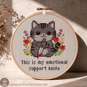Puede incluir: Un bastidor de bordado circular de madera con un patrón de punto de cruz de un gato atigrado gris sosteniendo un cuchillo, rodeado de flores coloridas. El texto debajo del gato dice "This is my emotional support knife" en letras negras.