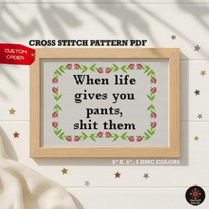 Puede incluir: Patrón de punto de cruz en un marco de madera. El texto dice: "When life gives you pants, shit them". El diseño está rodeado por un borde floral. Las dimensiones son 20 cm x 15 cm.