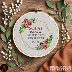 May include: A cross-stitch embroidery hoop with the text "SQUAT BECAUSE NO ONE RAPS ABOUT LITTLE BUTTS." The design includes floral and geometric elements in a wooden hoop. The background is a wooden surface.