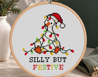 May include: A cross-stitch pattern in a wooden hoop featuring a white goose wearing a red Santa hat, tangled in colorful Christmas lights. The text below reads SILLY BUT FESTIVE in black and multicolored block letters on white fabric.