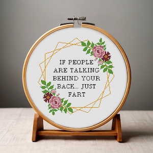 May include: A cross-stitch hoop with the text "IF PEOPLE ARE TALKING BEHIND YOUR BACK... JUST FART". The design includes a gold geometric shape and floral accents in pink, burgundy, and green. The hoop is displayed on a wooden stand.