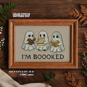 以下が含まれることがあります： 本を読む3体の幽霊のクロスステッチパターン。テキスト「I'M BOOOKED」が下にあります。フレームは木製です。パターンのサイズは21.59 cm x 13.97 cmで、7色のDMCカラーです。