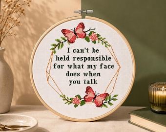 Puede incluir: Un bastidor de bordado circular de madera que contiene un patrón de punto de cruz sobre tela blanca. El diseño presenta dos mariposas rosas, hojas verdes y un marco geométrico que rodea el texto negro: I can't be held responsible for what my face does when you talk.