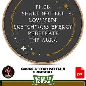 May include: A circular wooden embroidery hoop containing a cross-stitch design on black fabric. The text reads: THOU SHALT NOT LET LOW-VIBIN' SKETCHY-ASS ENERGY PENETRATE THY AURA. The design features a gold crescent moon and small white and gold stars.
