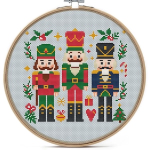 May include: A cross-stitch embroidery hoop featuring three toy soldier nutcrackers in red, green, and blue uniforms. The design includes Christmas-themed elements like holly, stars, and a candy cane, all set against a light blue background.