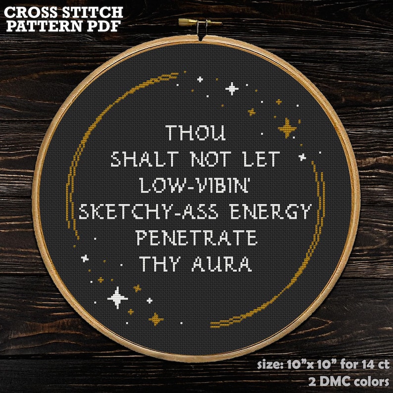 May include: A circular wooden embroidery hoop containing black fabric with white cross-stitched text that reads: THOU SHALT NOT LET LOW-VIBIN' SKETCHY-ASS ENERGY PENETRATE THY AURA. The design includes gold and white stitched stars and a curved line.