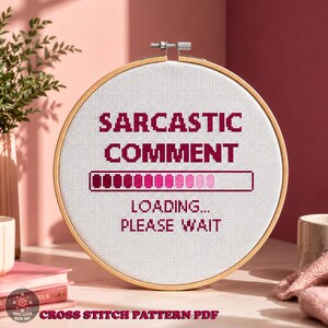 Pode incluir: Um bastidor de bordado circular de madeira contendo tecido Aida branco com um design em ponto cruz. O texto diz SARCASTIC COMMENT, seguido por uma barra de carregamento rosa com gradiente e o texto LOADING... PLEASE WAIT em letras rosa.