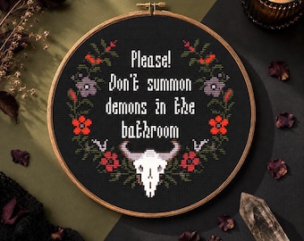 May include: A circular wooden embroidery hoop containing a black cross-stitch fabric with the white text "Please! Don't summon demons in the bathroom" surrounded by a floral wreath and a white bull skull at the bottom in red, green, and purple.