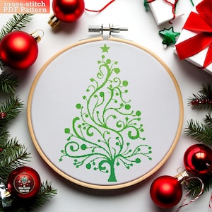 May include: A green and white cross-stitch pattern of a stylized Christmas tree in a wooden embroidery hoop. The hoop is surrounded by red Christmas ornaments and evergreen branches.