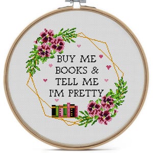 A bookish cross-stitch pattern with the text "BUY ME BOOKS & TELL ME I'M PRETTY". The design includes a floral wreath with pink flowers, green leaves, and a gold geometric shape. Small pink hearts and stacks of books are also included.